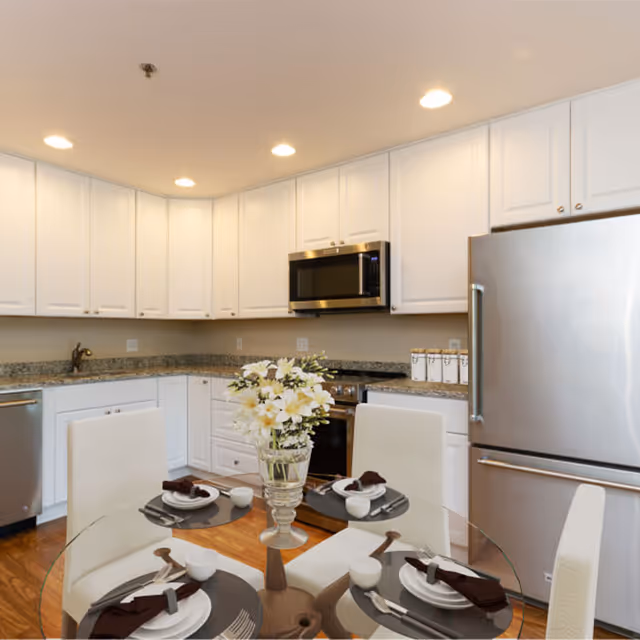 Bright modern kitchen with white cabinets, stainless steel appliances, and a glass dining table set for four with a floral centerpiece.