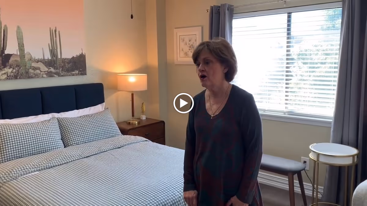 A bedroom with a neatly made bed featuring checkered bedding and a dark blue headboard. A woman stands near a window with blinds and gray curtains. There is a wooden nightstand with a lamp and decorative items, a bench under the window, and a small round side table. A framed picture hangs on the wall above the nightstand.