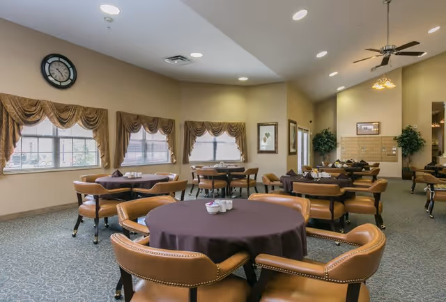 A spacious dining room with several round tables covered with dark tablecloths, surrounded by cushioned brown chairs. The room has large windows with decorative valances, a wall clock, framed artwork, and ceiling fans with lights. The carpet is patterned and the walls are painted a light beige color.