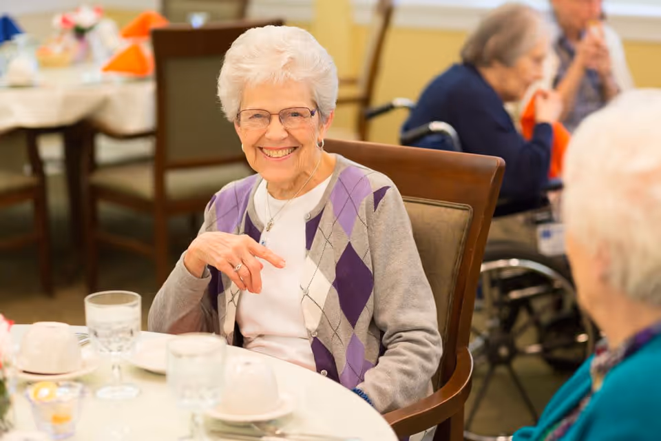 An elderly woman with white hair and glasses smiling while sitting at a dining table in a senior living facility. She is wearing a white shirt and a gray cardigan with purple and white diamond patterns. Other elderly individuals are visible in the background, some seated in wheelchairs, in a warmly lit dining area.