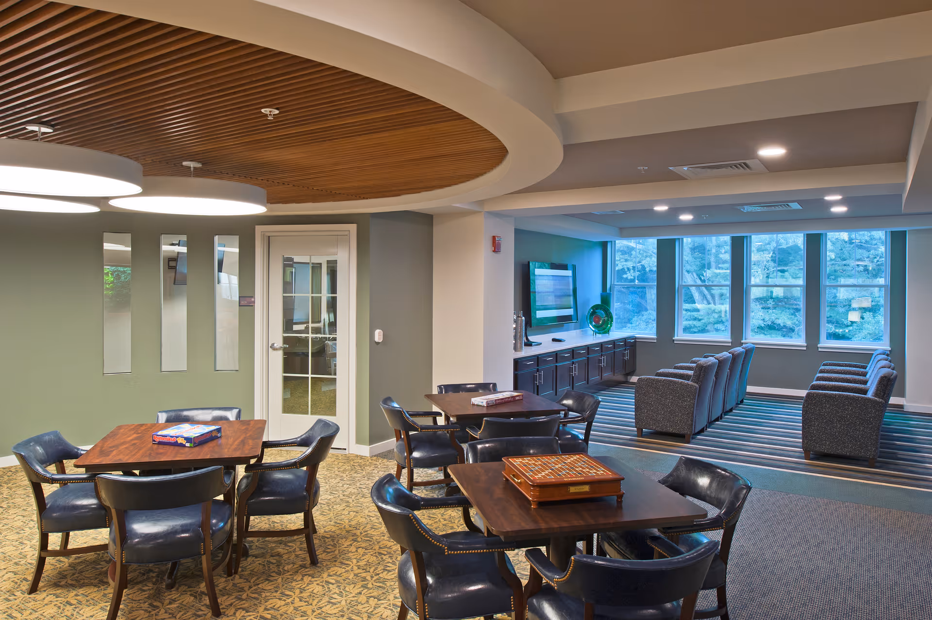 A spacious common area in a senior living facility featuring multiple tables with chairs arranged for games, including a Scrabble board and a Rummikub game box. The room has a modern design with a wooden slatted ceiling feature, green walls, and large windows letting in natural light. There is a seating area with armchairs facing a wall-mounted television and a long cabinet beneath it.