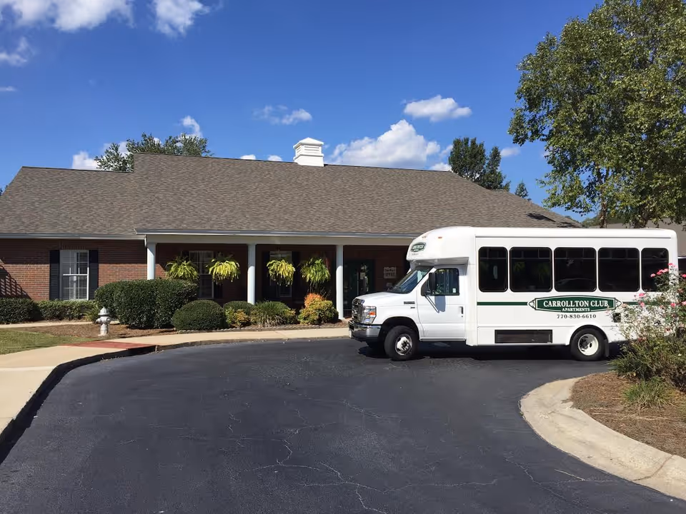 Exterior view of a single-story brick building with a gray shingled roof, surrounded by green bushes and hanging plants. A white shuttle bus with the sign 'Carrollton Club Apartments' and a phone number is parked on the driveway in front of the building. The sky is blue with scattered clouds.