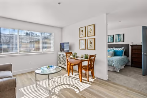 A bright and airy living area with a large window letting in natural light. The room features a small wooden dining table with two chairs, a round glass coffee table, and a TV on a white cabinet. Adjacent to the living area is a bedroom with a bed dressed in blue and white bedding, a wooden dresser, and artwork on the walls.