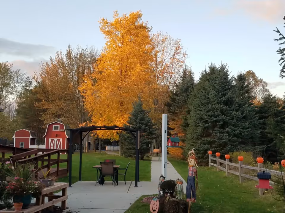 Outdoor garden area with a concrete pathway, a pergola with a table and chairs underneath, surrounded by green grass and trees with autumn foliage. There are small red barn-like structures on the left, a wooden fence decorated with small pumpkin ornaments on the right, and scarecrow decorations near the pathway.
