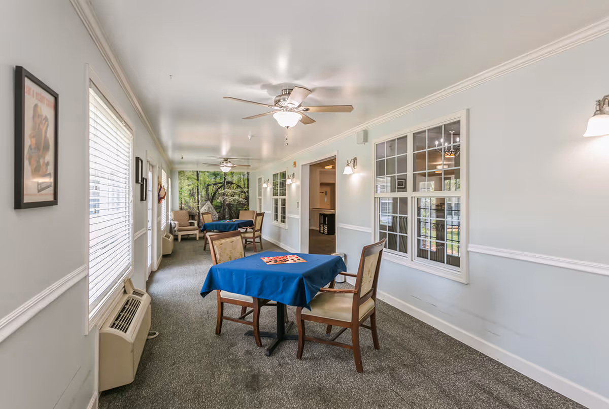 A bright, narrow common area in a senior living facility with tables covered in blue tablecloths and chairs arranged around them. The room has ceiling fans with lights, large windows with white blinds on one side, and a wall with windows looking into another room. There are framed pictures on the walls and a nature-themed mural at the far end of the room.