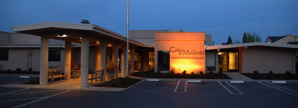 Exterior view of Casa de Modesto senior living facility at dusk, showing the entrance with columns, benches, and illuminated signage.