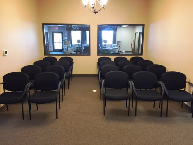 A small meeting or conference room with four rows of black chairs arranged in two columns facing forward. The room has beige walls and carpeted floor. Two large windows on the far wall look into another room with sofas and lamps. A chandelier hangs from the ceiling.