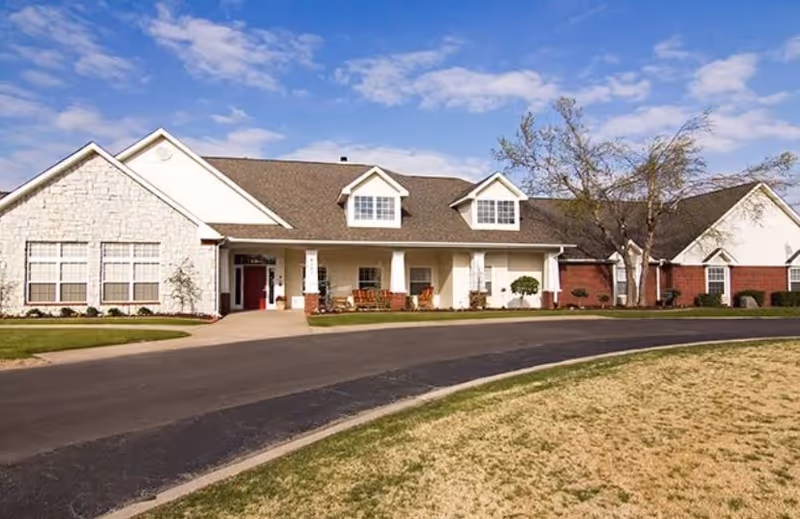 Single-story assisted living building with a covered front porch, dormer windows, and a circular driveway under a blue sky.