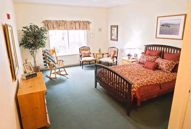 A furnished bedroom with a double bed, nightstand, dresser, rocking chair and a seating area by a large window.
