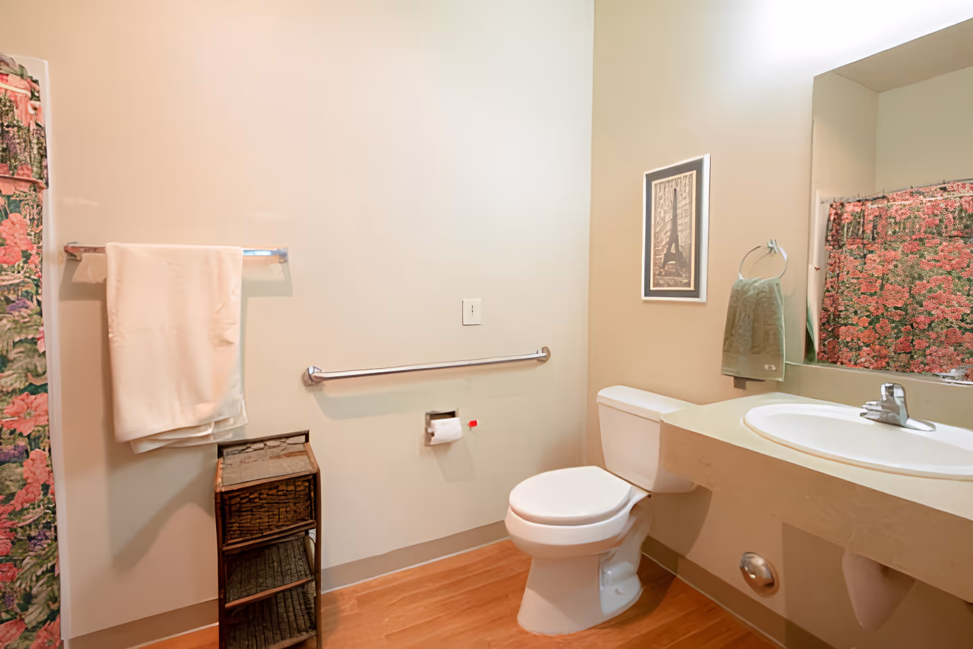 A clean bathroom with a white toilet, a sink with a faucet, a large mirror, and a floral shower curtain. There is a towel rack with a white towel, a small wicker storage unit, a green hand towel hanging on a ring, and a framed picture on the wall.