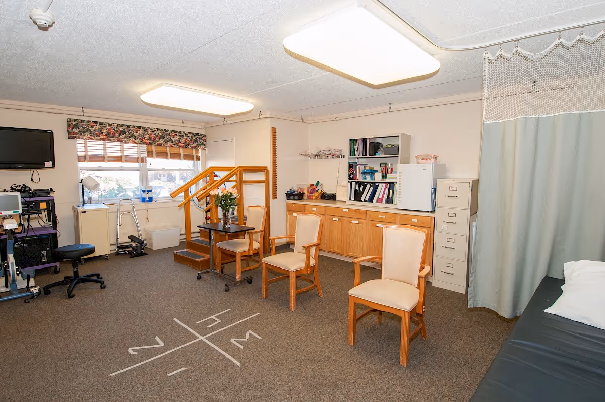 A therapy or rehabilitation room with exercise equipment, a small set of wooden stairs with a handrail, three beige chairs around a small table with flowers, a filing cabinet, shelves with binders and supplies, a curtain partially covering a padded examination or therapy table, and a window with floral valance letting in natural light.