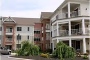 Front exterior of a multi-story senior living building with balconies, brick accents, a parked vehicle, and landscaped shrubs.