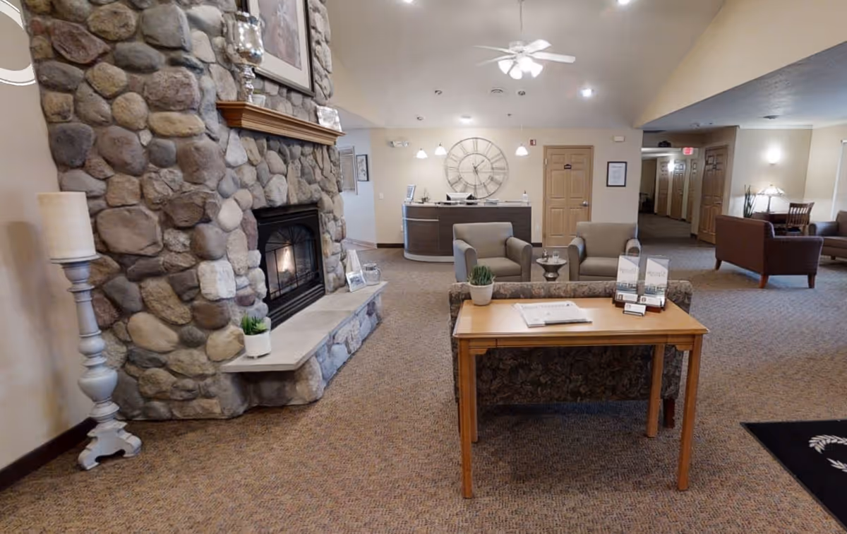 Interior view of a senior living facility lounge area featuring a stone fireplace with a lit fire, a wooden mantel with decorative items, a tall candle holder, a wooden table with brochures and a small plant, two armchairs, and a reception desk with a large wall clock behind it. The space is carpeted and well-lit with ceiling lights.