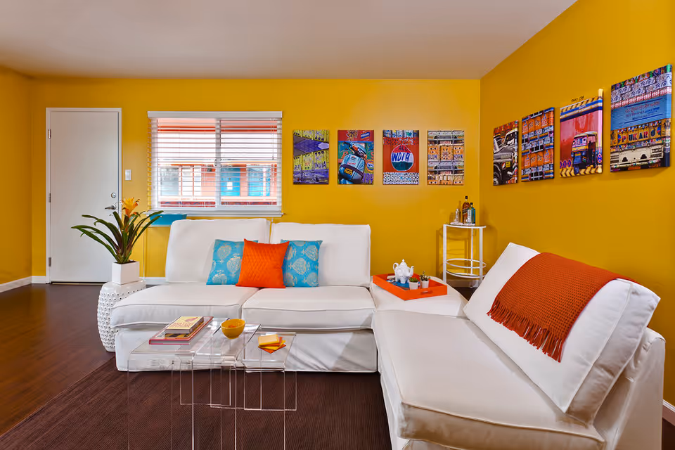 A bright living room with yellow walls featuring a white sectional sofa adorned with blue and orange pillows and an orange throw blanket. A clear nested coffee table holds books and a bowl. Colorful artwork hangs on the walls, and a small white side table with bottles is in the corner. A window with blinds and a white door are visible in the background.