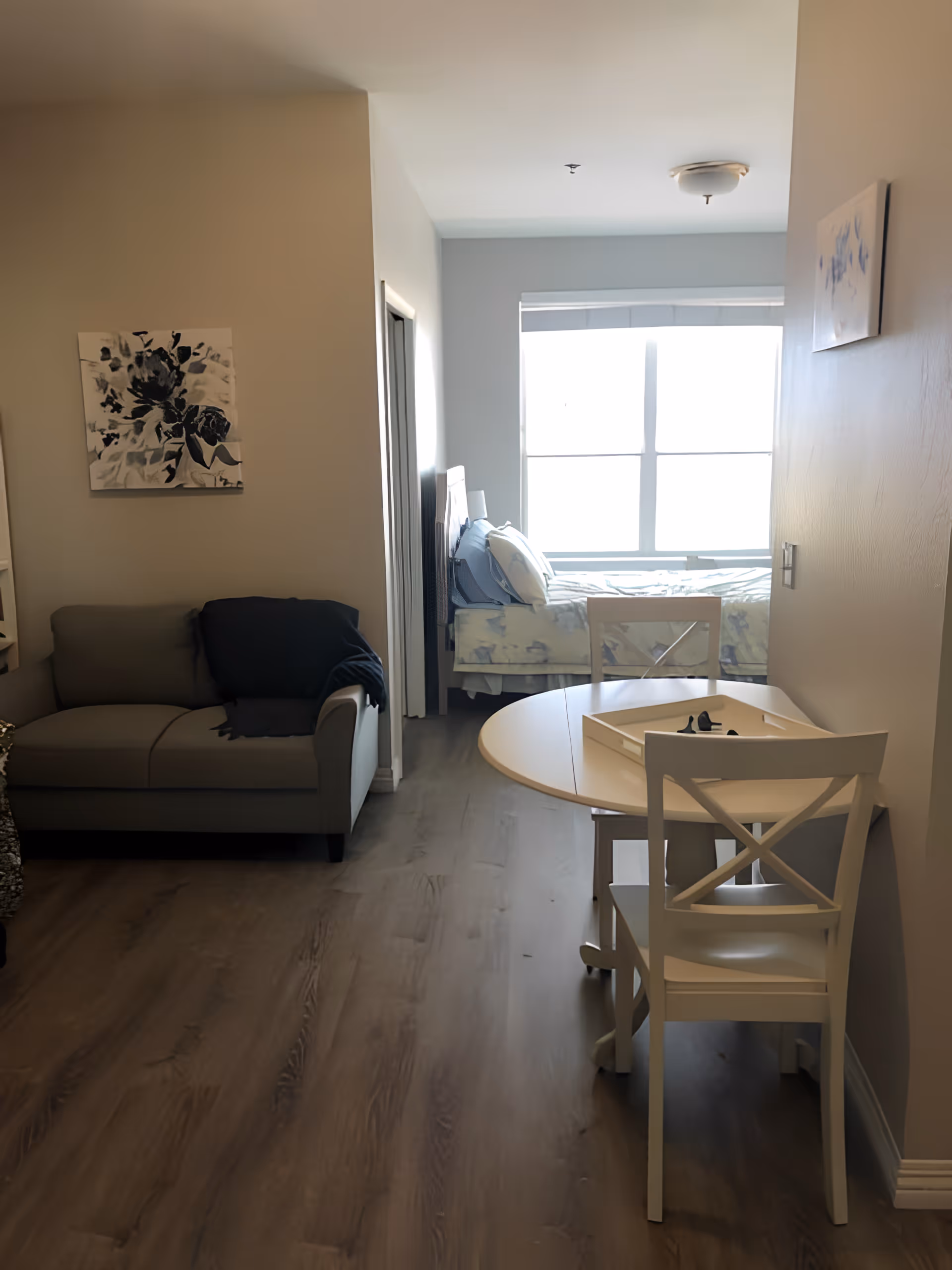 Interior view of a senior living facility room with a small round dining table and two white chairs in the foreground, a gray couch with a dark blanket on the left, and a bed near a large window in the background. The room has light-colored walls and wood flooring, with two pieces of wall art visible.