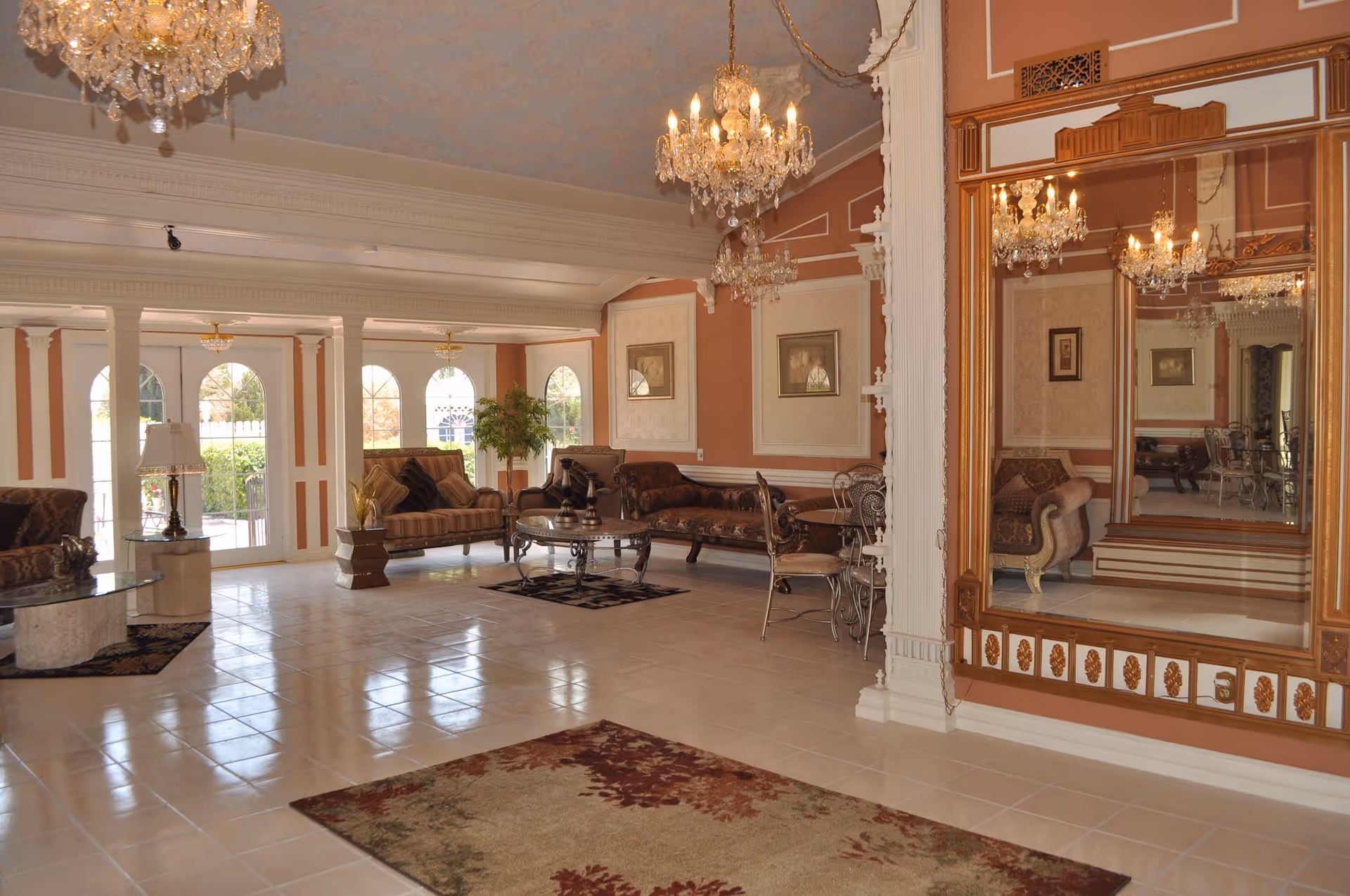 A spacious and elegant living room with large windows and glass doors letting in natural light. The room features ornate chandeliers, classic upholstered sofas, a glass coffee table, decorative rugs on tiled floors, framed artwork on the walls, and a large decorative mirror reflecting the room's interior.