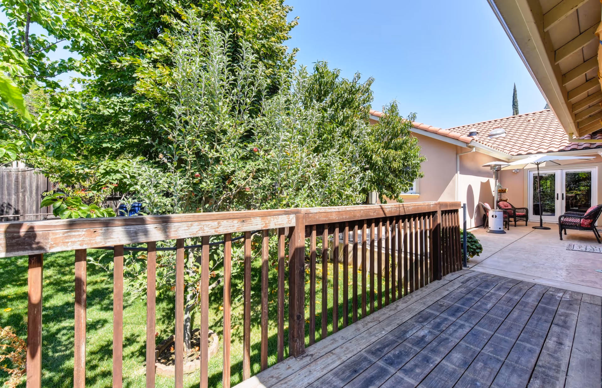 Wooden deck and railing overlooking a green backyard with trees and a patio seating area next to a single-story house.
