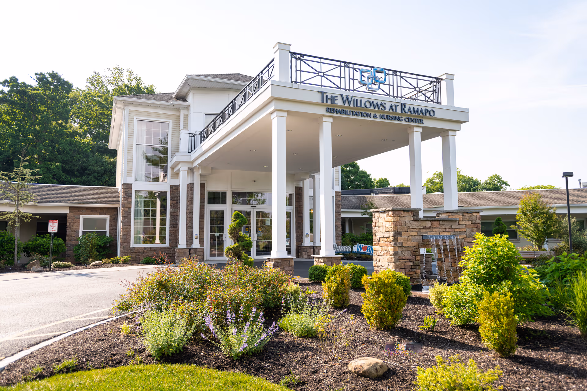 Front exterior view of The Willows at Ramapo Rehabilitation & Nursing Center building with a covered entrance supported by white columns, landscaped garden with bushes and flowers, and a stone water feature in front.