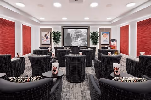A cozy movie theater room with multiple rows of black cushioned chairs facing a large screen. The room has red window shades, two potted plants on either side of the screen, and movie posters on the walls. There is also a popcorn machine and popcorn containers on small tables beside the chairs.