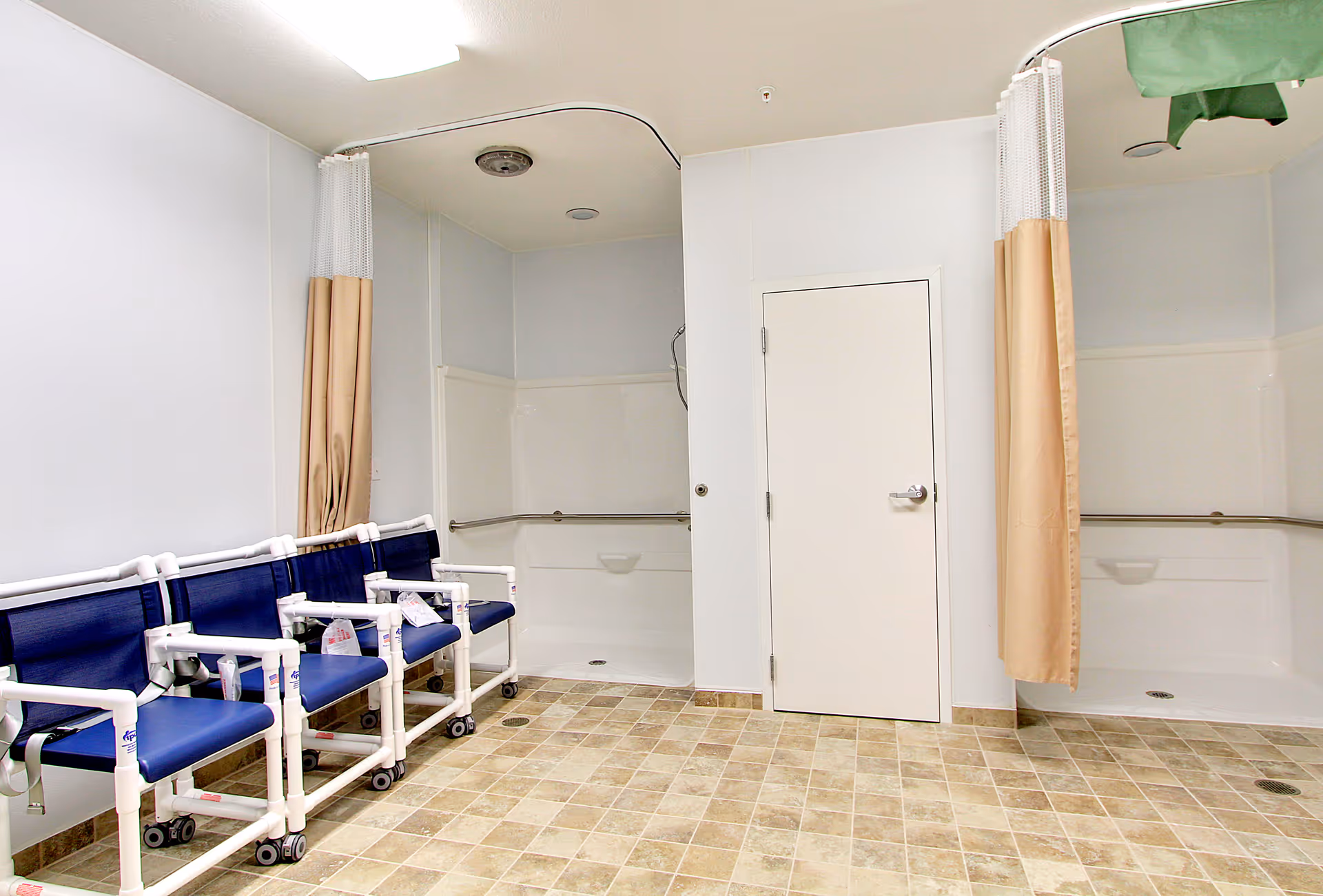A clean, accessible shower room with two shower stalls separated by beige privacy curtains. There are four blue and white shower chairs lined up against the left wall. The floor is tiled with a beige and light brown pattern, and there is a white door in the center of the back wall.