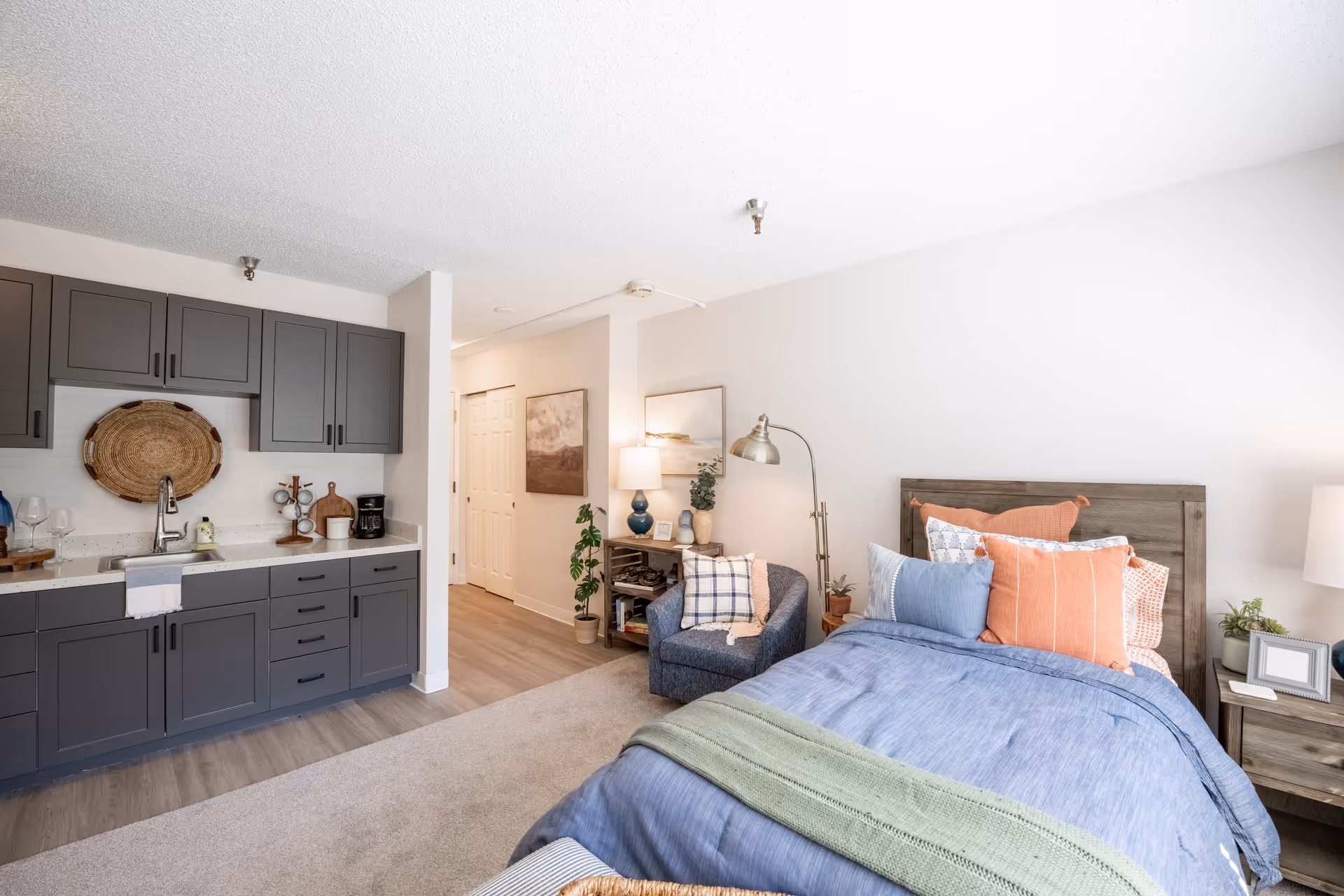 A bright and cozy senior living studio apartment featuring a bed with blue and orange pillows and a green throw blanket. Next to the bed is a wooden nightstand with a lamp and small plants. There is a comfortable blue armchair with a plaid pillow and a small wooden shelf with decorative items and a lamp. The kitchenette has gray cabinets, a sink, a coffee maker, and decorative items on the countertop. The floor is a combination of carpet and wood, and the walls are painted white with framed artwork.