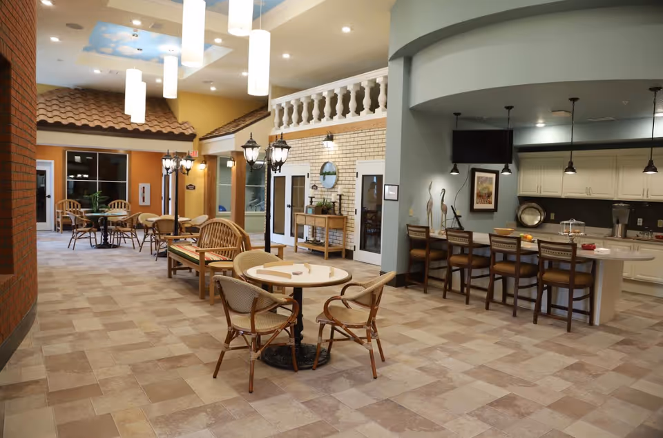 Bright communal dining and lounge area with round tables, wicker chairs, a breakfast bar and kitchenette in a senior living facility.