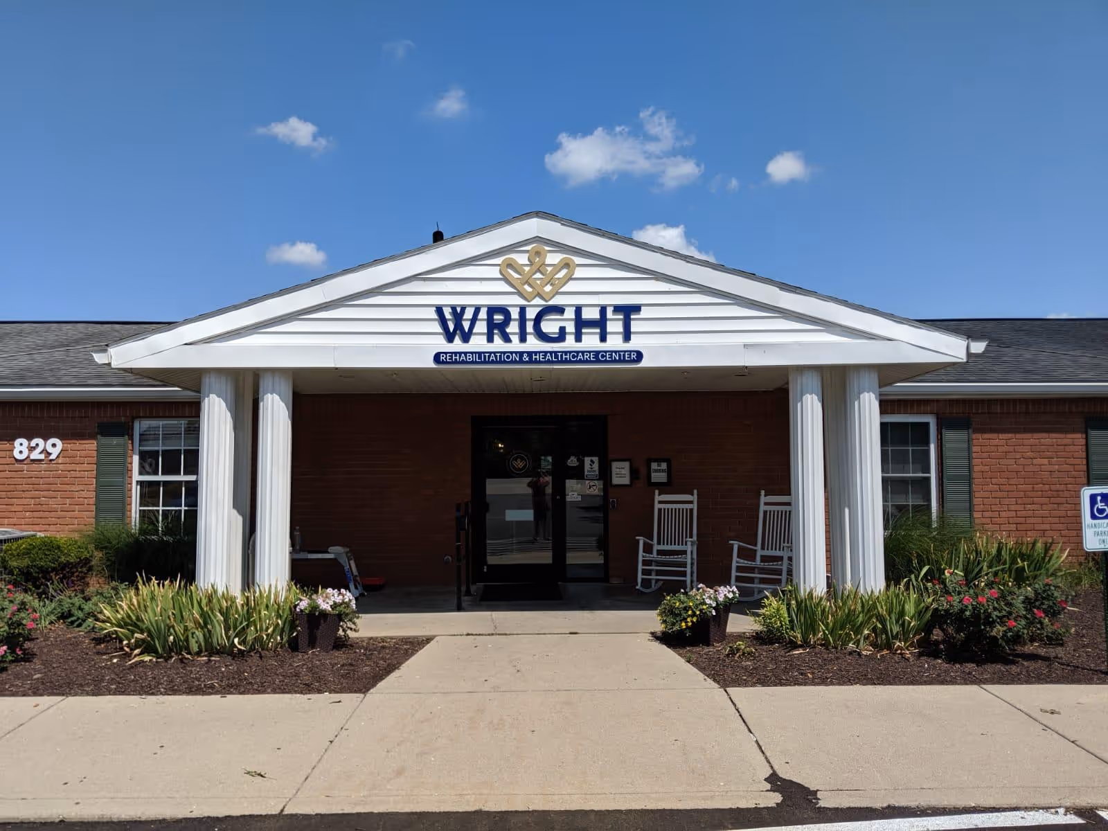 Front exterior view of Wright Rehabilitation and Healthcare Center building with a white portico supported by columns, two white rocking chairs on the porch, flower beds on either side of the entrance, and a clear blue sky with a few clouds.