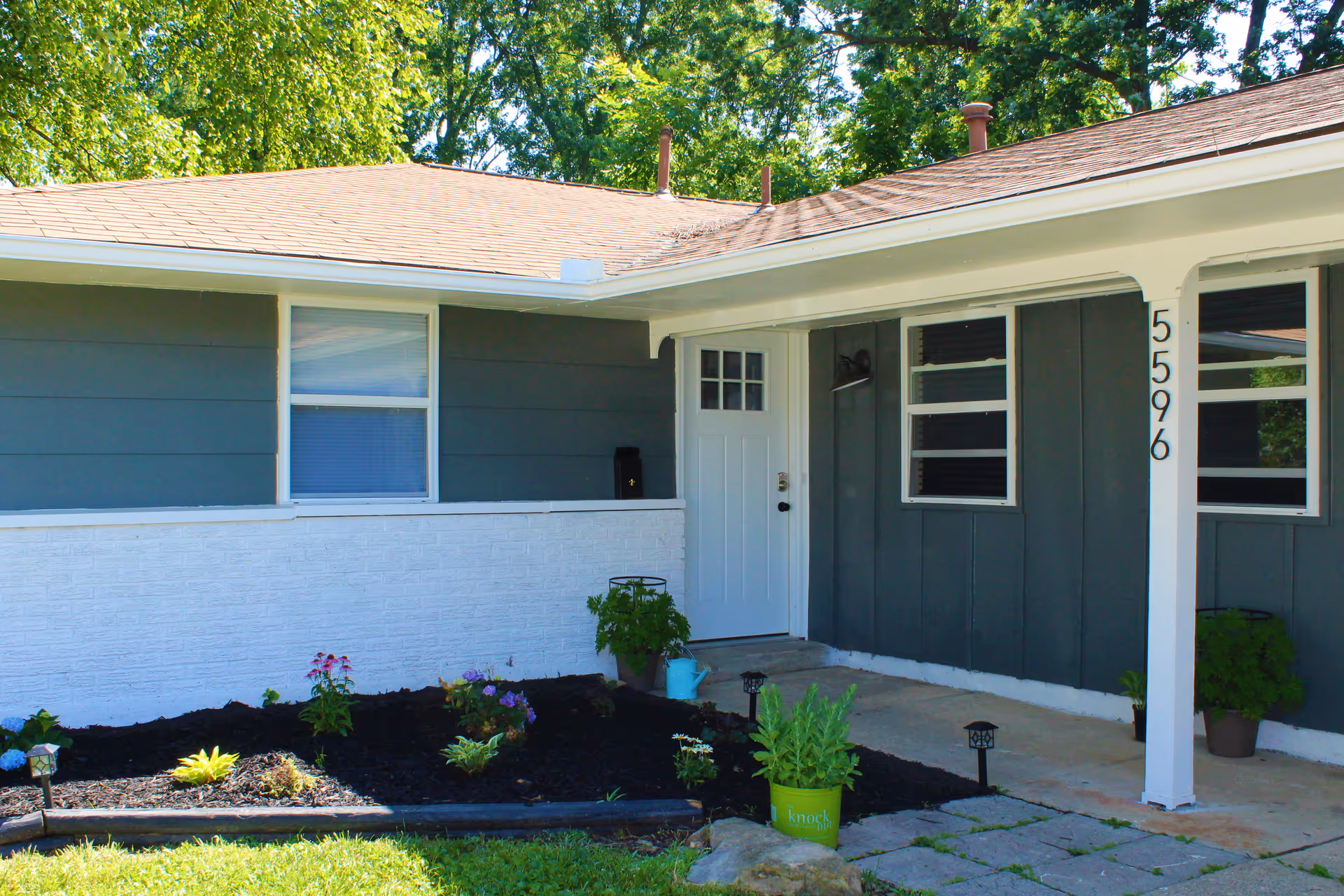 Front entrance of a single-story house with a white door, dark gray siding, house number 5596, and a small garden bed.