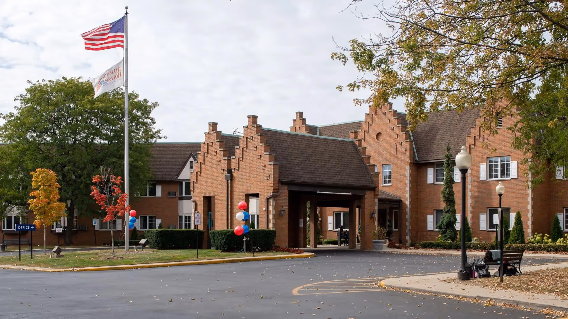 Exterior view of a brick senior living facility building with a covered entrance. There are two flagpoles with the American flag and another flag, trees with autumn foliage, and a paved driveway. Benches and lamp posts are visible near the entrance.