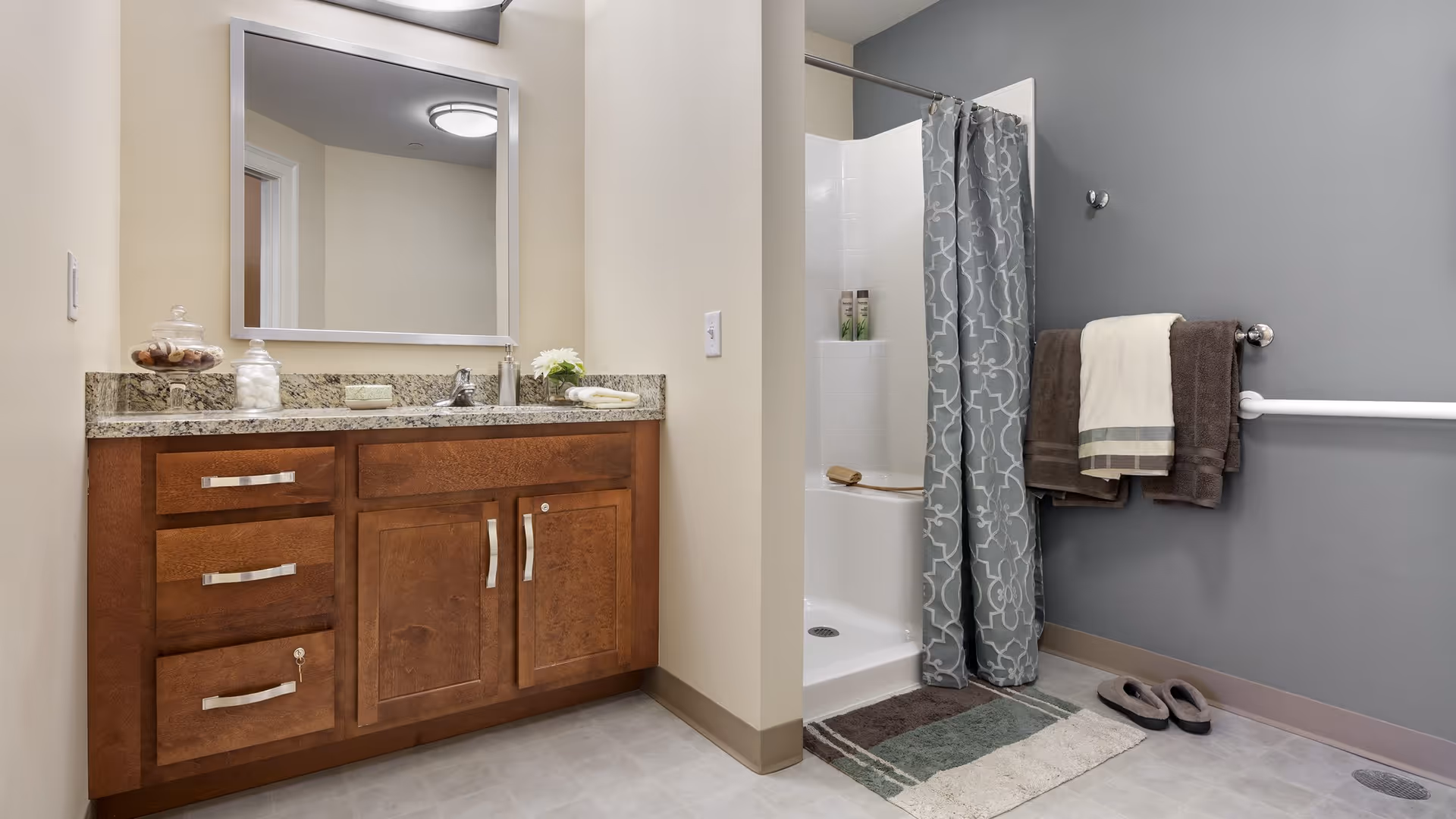 A clean and modern bathroom featuring a wooden vanity with granite countertop, a large mirror above the sink, and a walk-in shower with a patterned gray shower curtain. Towels hang on a rack mounted on a gray wall, and a pair of slippers are placed on the floor next to a bath mat.