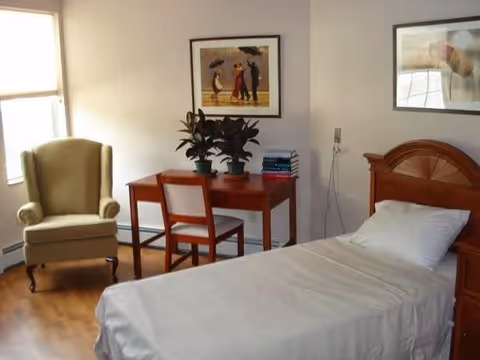 A simple bedroom with a single bed covered in white bedding, a wooden headboard, a wooden desk with a chair, two potted plants, and a stack of books. There is a beige armchair near a window with blinds, and two framed pictures hanging on the light-colored walls.