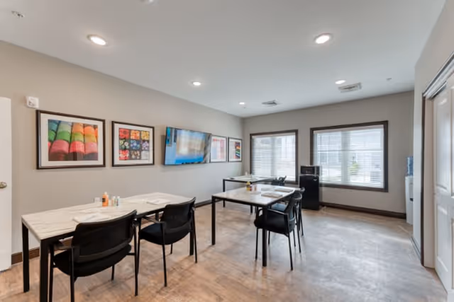 A bright room with two rectangular tables, each surrounded by black chairs. The tables have placemats and condiments on them. The walls are light gray with colorful framed artwork and a mounted flat-screen TV. Two large windows with blinds let in natural light. The floor is a light wood finish, and the ceiling has recessed lighting.