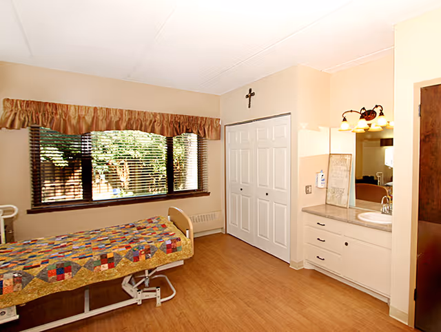 A well-lit bedroom in a senior living facility featuring a hospital-style bed with a colorful patchwork quilt, a large window with brown blinds and a valance, a white double-door closet, a wall-mounted cross above the closet, and a vanity area with a sink, mirror, and light fixture.