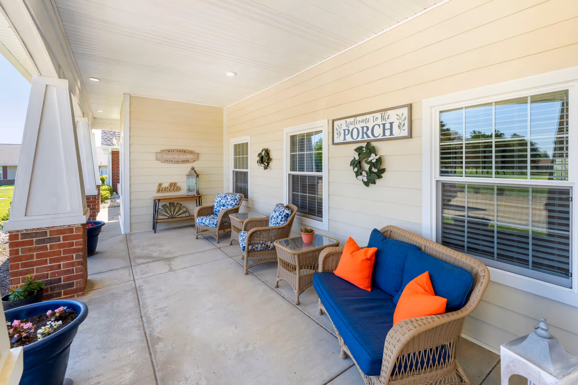 A covered porch area with beige siding and brick pillars. The porch is furnished with wicker furniture including a blue cushioned sofa with orange pillows, two armchairs with blue floral cushions, and two glass-top side tables. There are decorative wreaths on the wall and signs that read 'Welcome to the PORCH' and 'hello'. Several potted plants are placed along the edge of the porch.