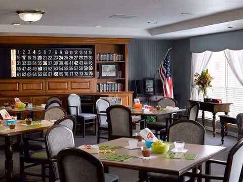A well-lit common dining area with several tables and chairs arranged neatly. Each table has placemats, cups, and snacks. In the background, there is a wooden bookshelf with books and a large bingo board mounted on the wall. An American flag stands in the corner near a window with white curtains, and a small table with a flower arrangement is placed nearby.