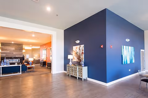 Interior view of a senior living facility showing a spacious common area with wooden flooring, a dark blue accent wall with two paintings, a decorative console table with a lamp and vases, and an open area leading to a lounge with seating and a fireplace.