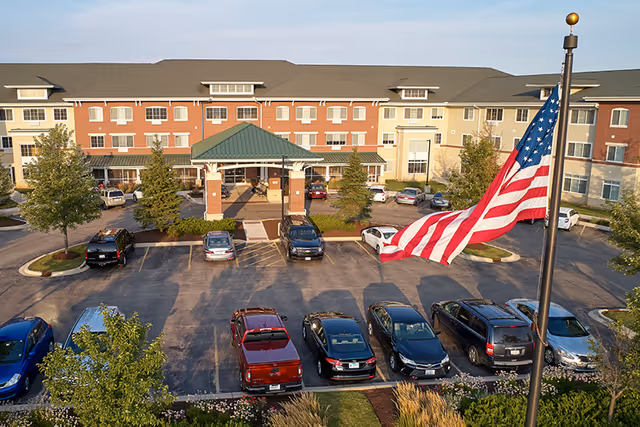 Front exterior of a multi-story senior living building with a covered entrance, parking lot, and an American flag in the foreground.
