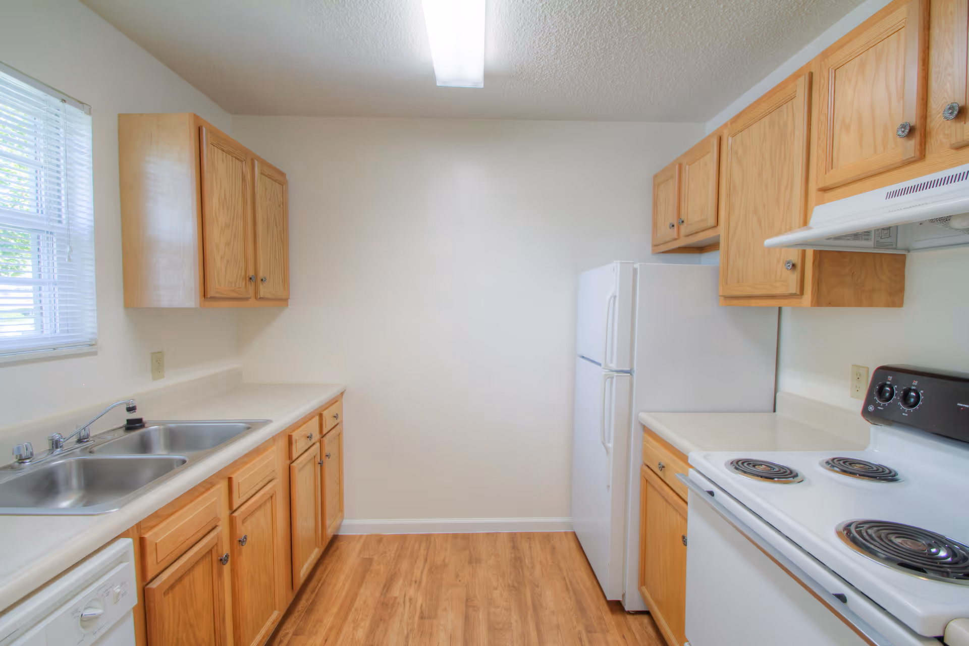A clean kitchen with wooden cabinets, a double stainless steel sink, a white electric stove with four burners, a white refrigerator, and a window with blinds letting in natural light. The floor is wooden and the walls are painted white.