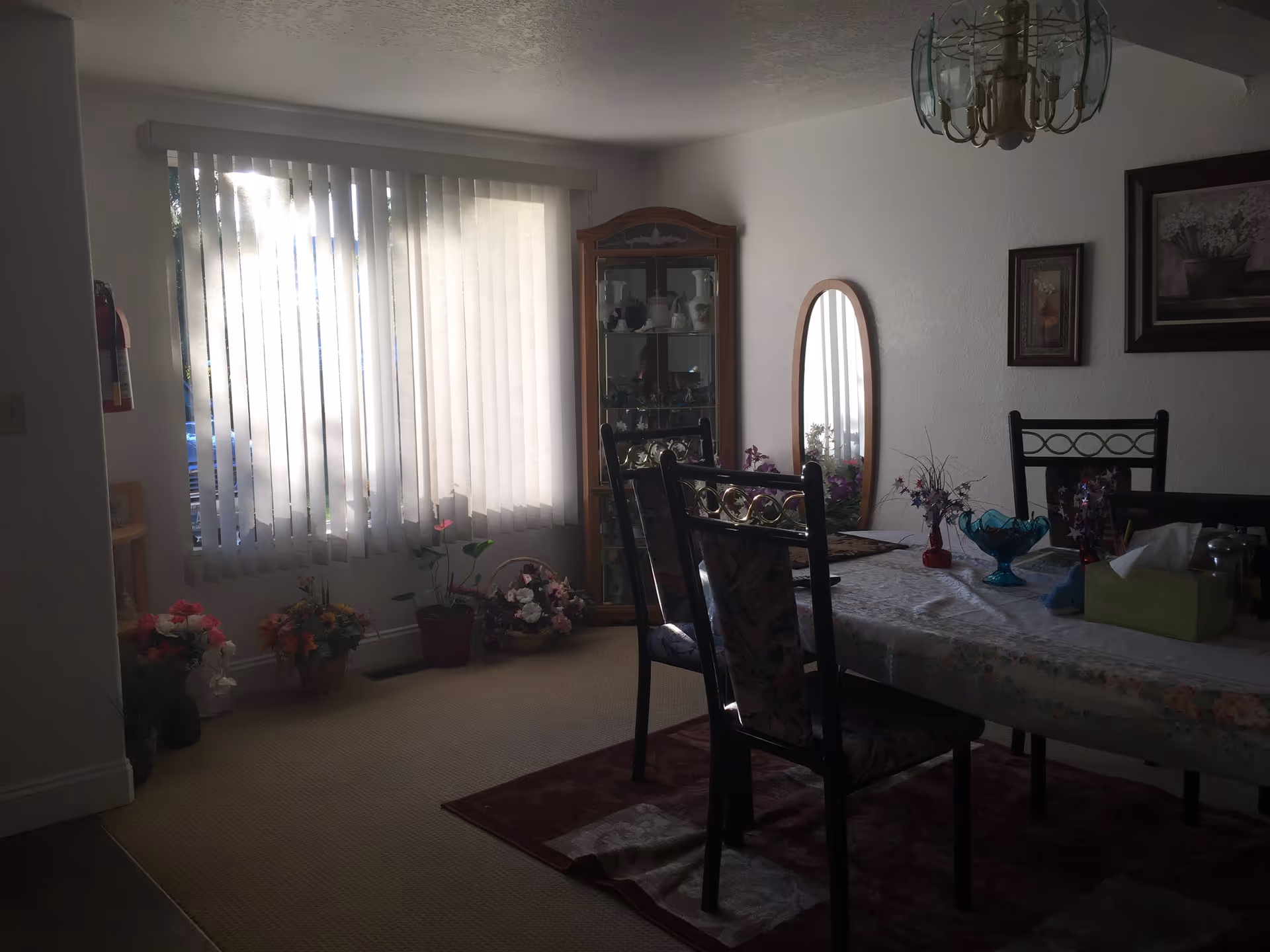 A dining room with a table covered by a floral tablecloth and surrounded by chairs. The room has a large window with vertical blinds letting in sunlight. There are several flower arrangements on the floor near the window and decorative items on the table. A glass-fronted cabinet with dishes and a tall oval mirror are against the wall. Two framed pictures hang on the wall, and a chandelier is visible on the ceiling.