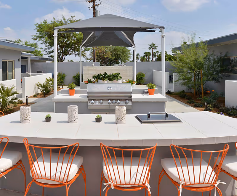 Outdoor patio area with a built-in stainless steel grill under a canopy, surrounded by a white tiled counter with decorative candle holders and small potted plants. Orange metal chairs with white cushions are arranged along the counter. The area is landscaped with small trees and plants, and there are buildings visible in the background under a partly cloudy sky.