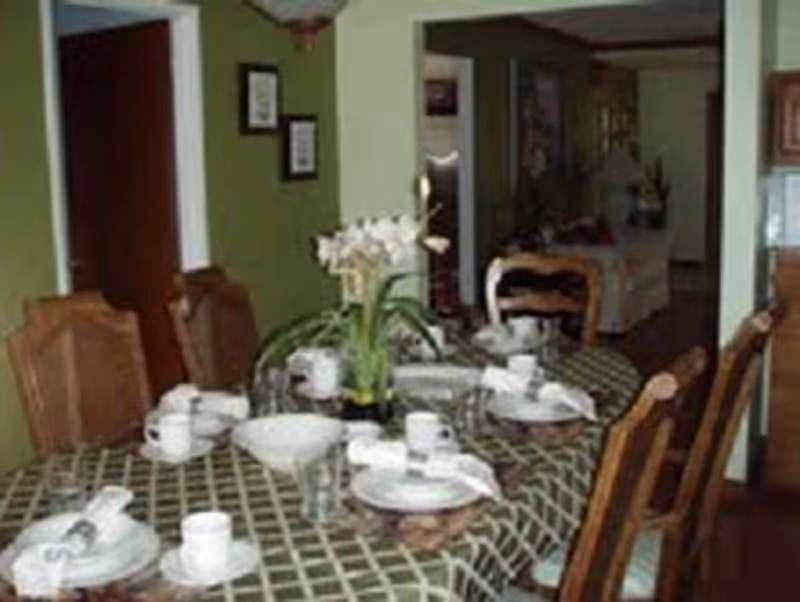 Dining room with a round table set for a meal with dishes, cups and a floral centerpiece, surrounded by wooden chairs.