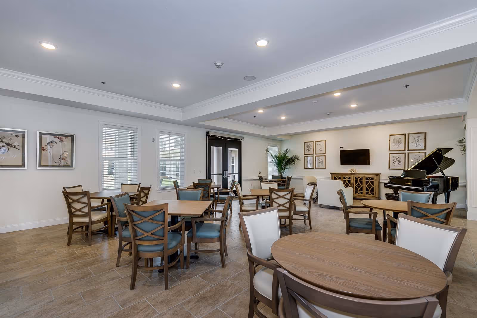 Bright communal dining and lounge area with round wooden tables and chairs, a grand piano, and a wall-mounted TV.
