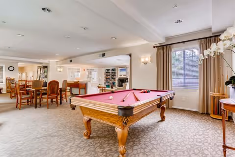 Communal activity room with a pink-felt pool table, seating and dining tables, bookshelves, and a large window with curtains.