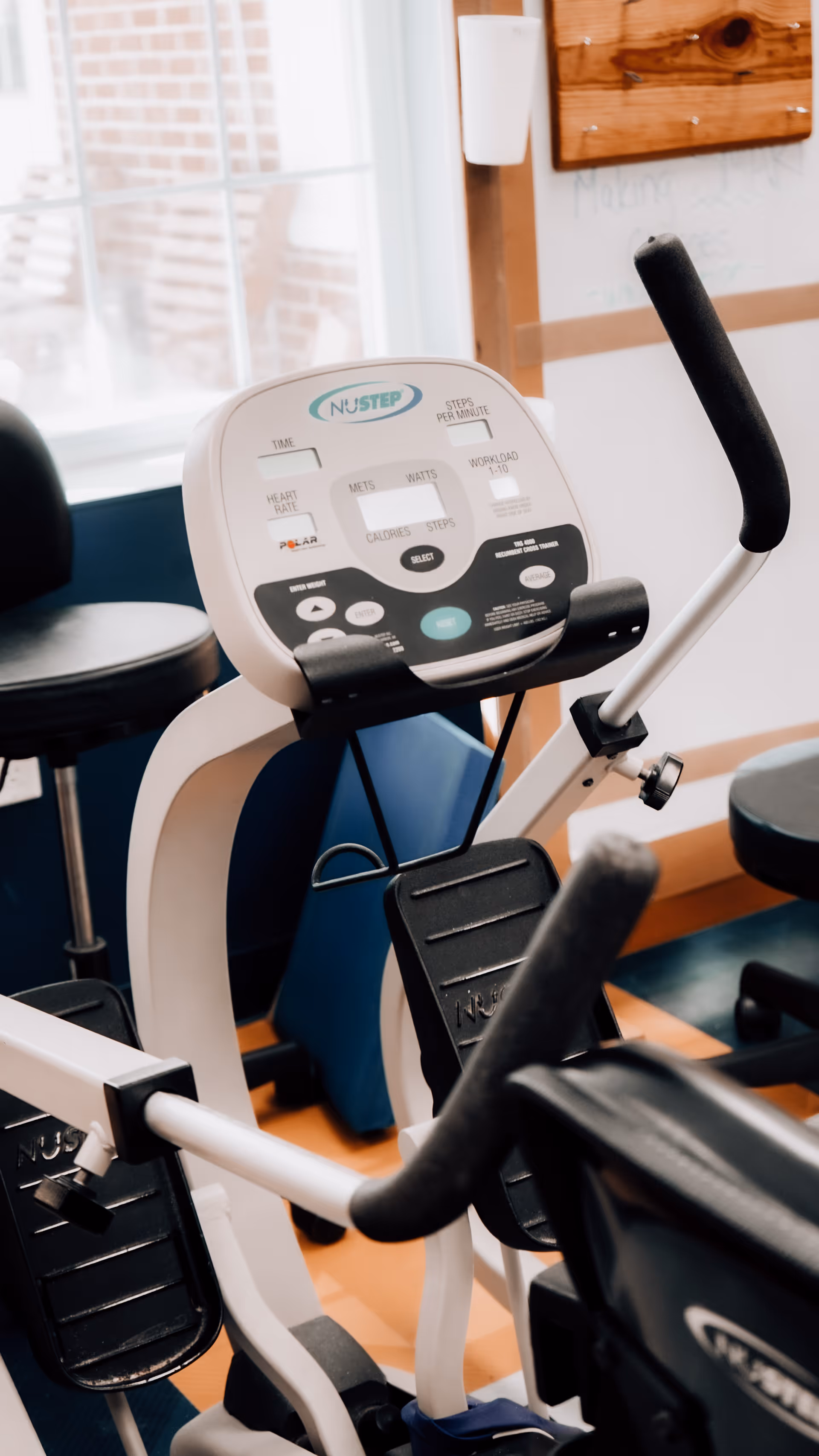 Close-up view of a NuStep exercise machine in a fitness or rehabilitation room with stools and a window in the background.