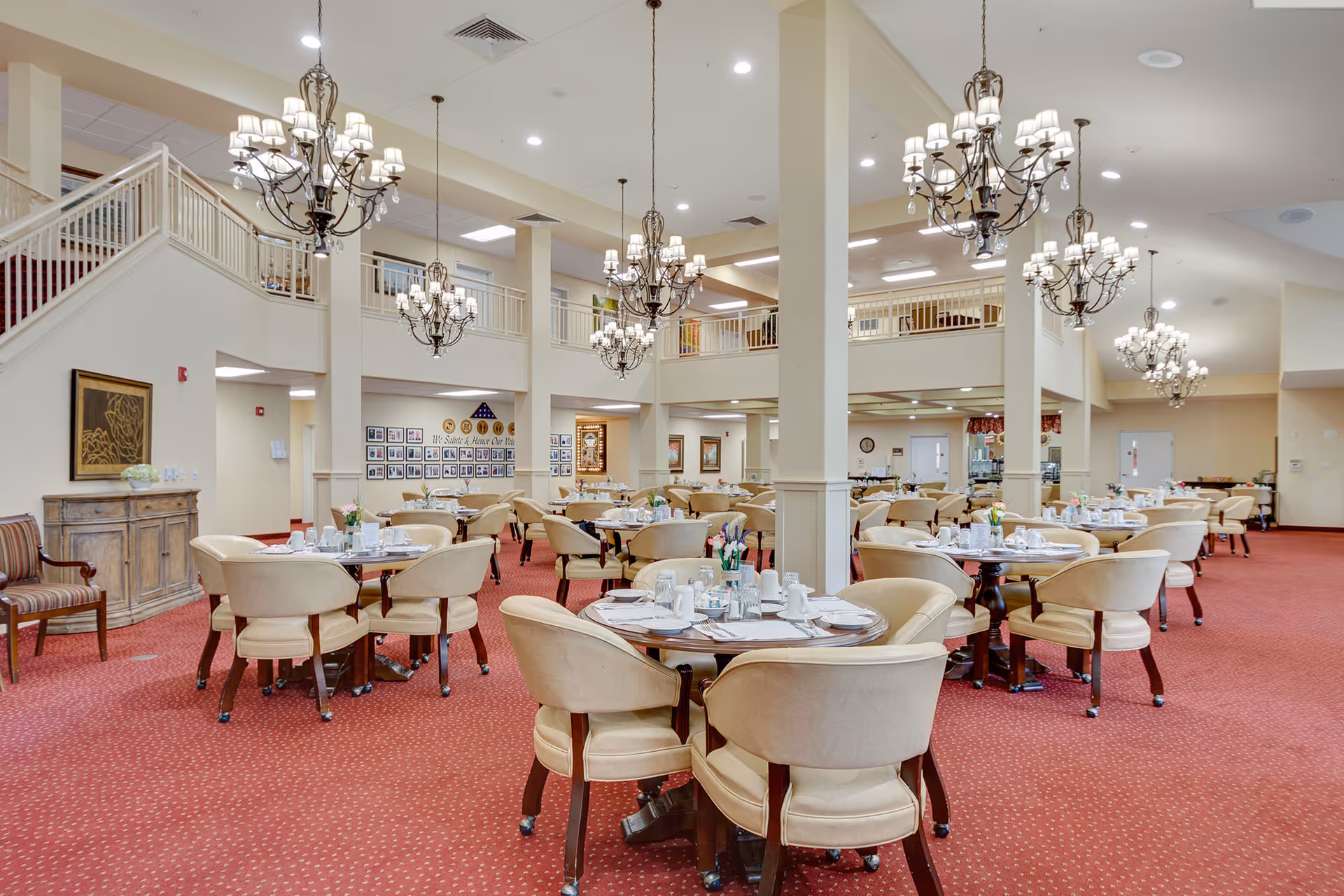 Spacious dining room with round tables, upholstered chairs, chandeliers, and red carpeting in a senior living facility.