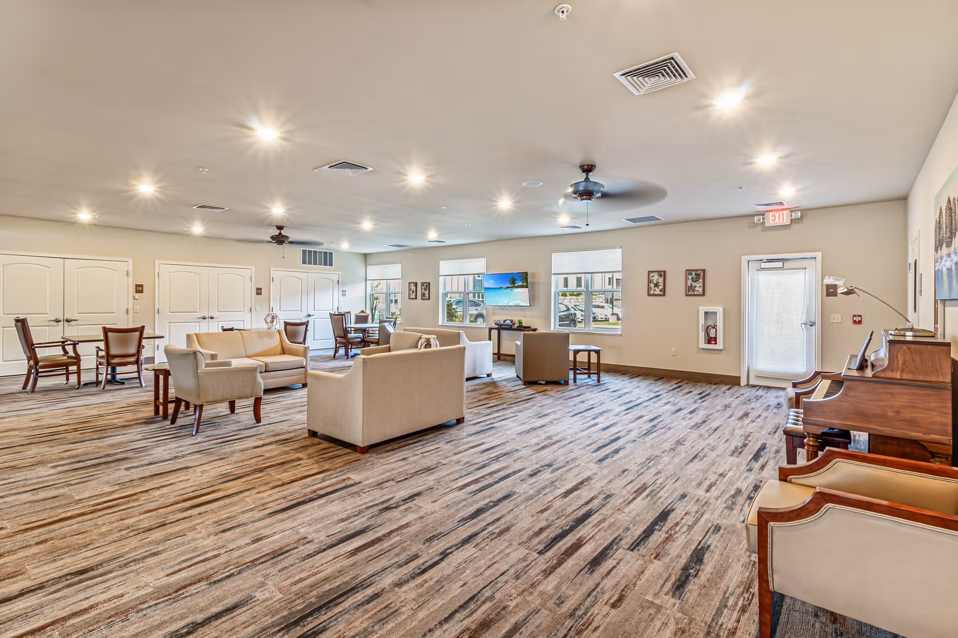 A spacious and well-lit common area in an assisted living facility featuring multiple seating arrangements with sofas and chairs, a piano on the right side, a wall-mounted TV, large windows letting in natural light, ceiling fans, and neutral-colored walls and carpet.