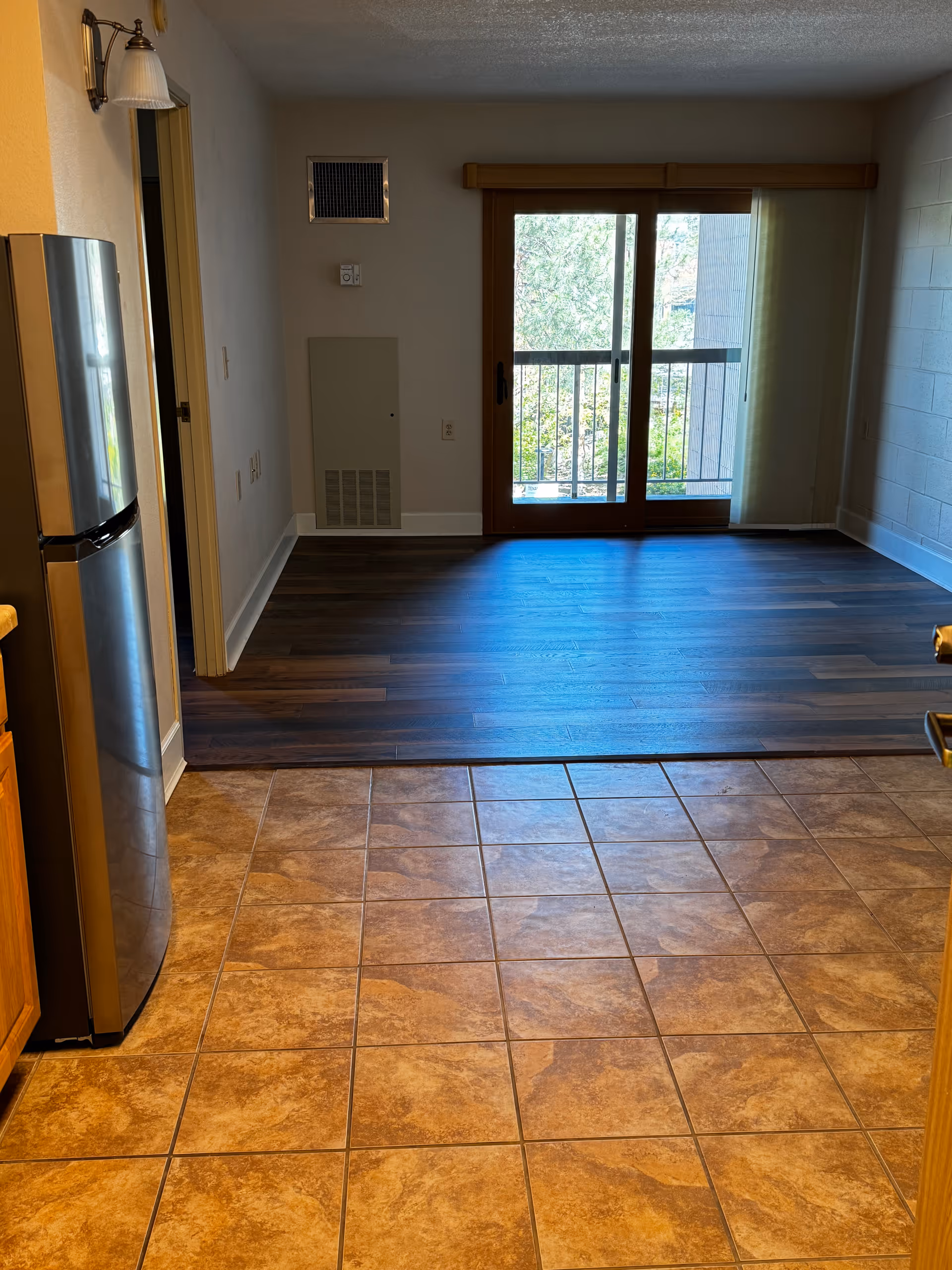 View from a kitchen area with tiled flooring looking into an empty living space with dark wood flooring and a sliding glass door leading to a small balcony with a railing and greenery outside. A stainless steel refrigerator is visible on the left side, and there is a wall vent and light fixture on the left wall.