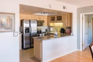 A modern kitchen with light wood cabinets, stainless steel refrigerator, microwave, and oven. The kitchen features a granite countertop island with a small decorative item on it. The walls are light-colored, and there is a framed painting on the left wall. The kitchen opens into a hallway and a dining area with wooden flooring.