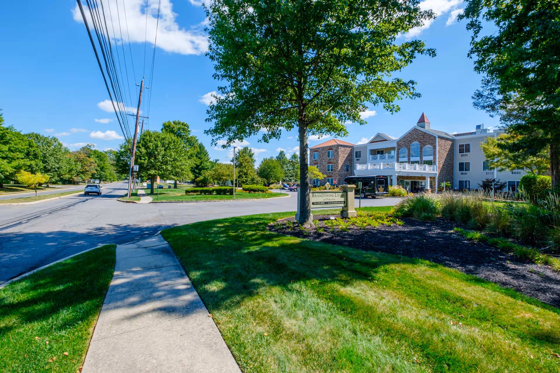 View of the exterior of Brandywine Princeton by Monarch, a multi-story senior living facility with a stone and beige facade, surrounded by green lawns, trees, and a clear blue sky. A sidewalk leads towards the building, and a sign with the facility's name is visible near the entrance.