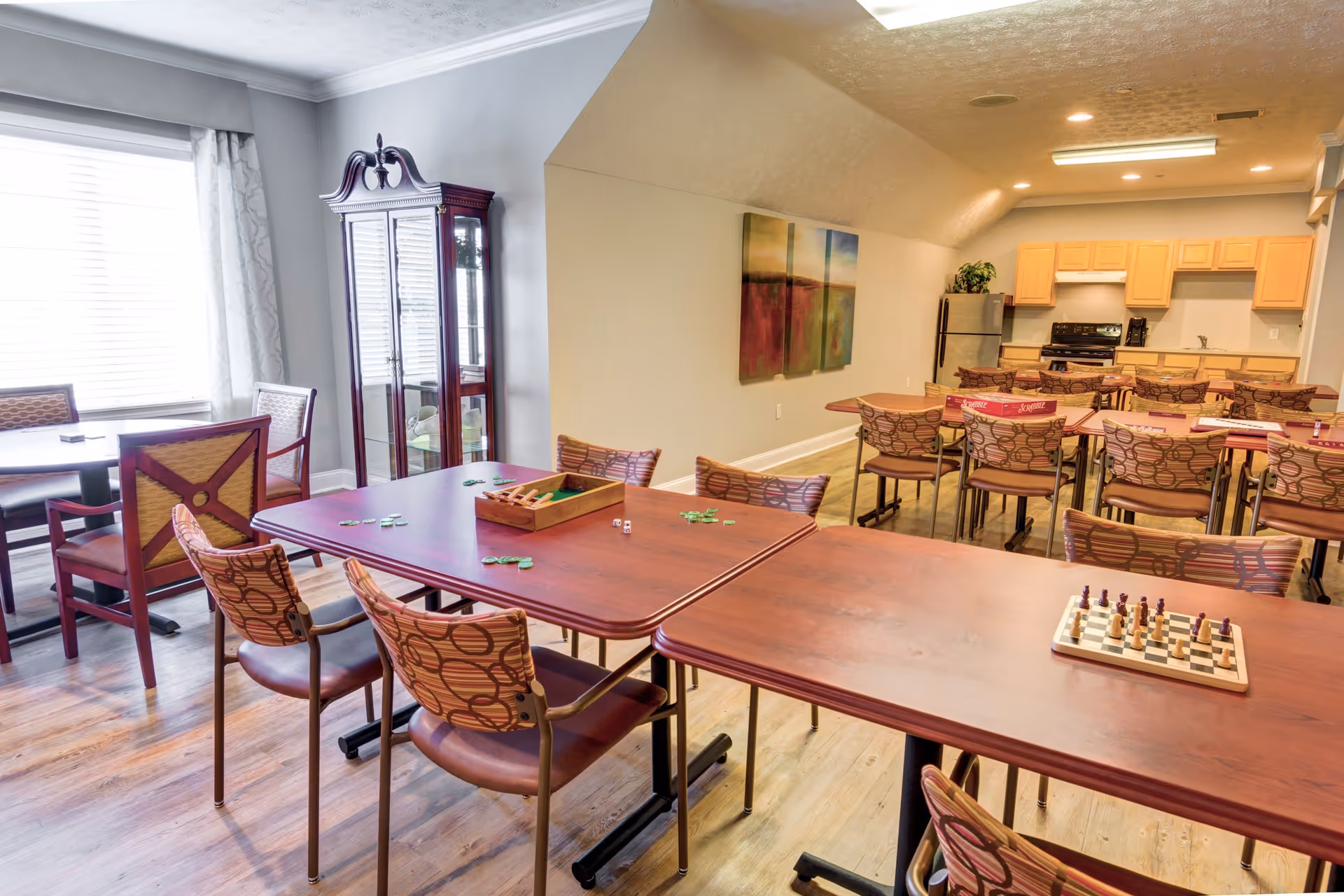 A bright activity room with multiple tables and chairs arranged for games and socializing. On the tables are game pieces, including a chessboard and other game tokens. The room has wooden flooring, a large window with curtains, a glass cabinet, and a kitchen area with light wood cabinets and appliances in the background. A colorful abstract painting hangs on the wall.